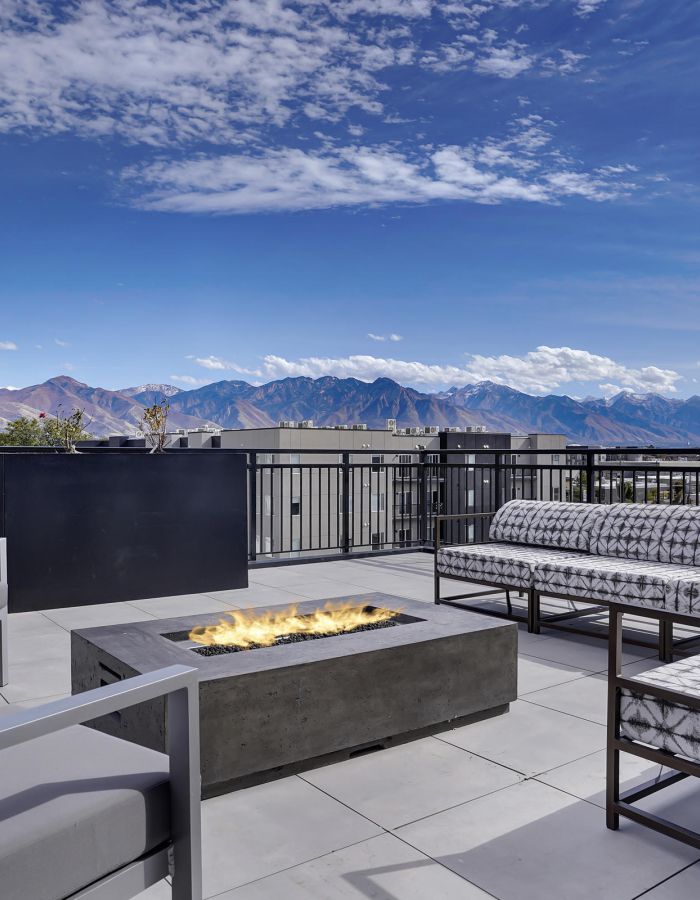 Modern rooftop patio with a fire pit, outdoor seating, and mountain views under a blue sky.