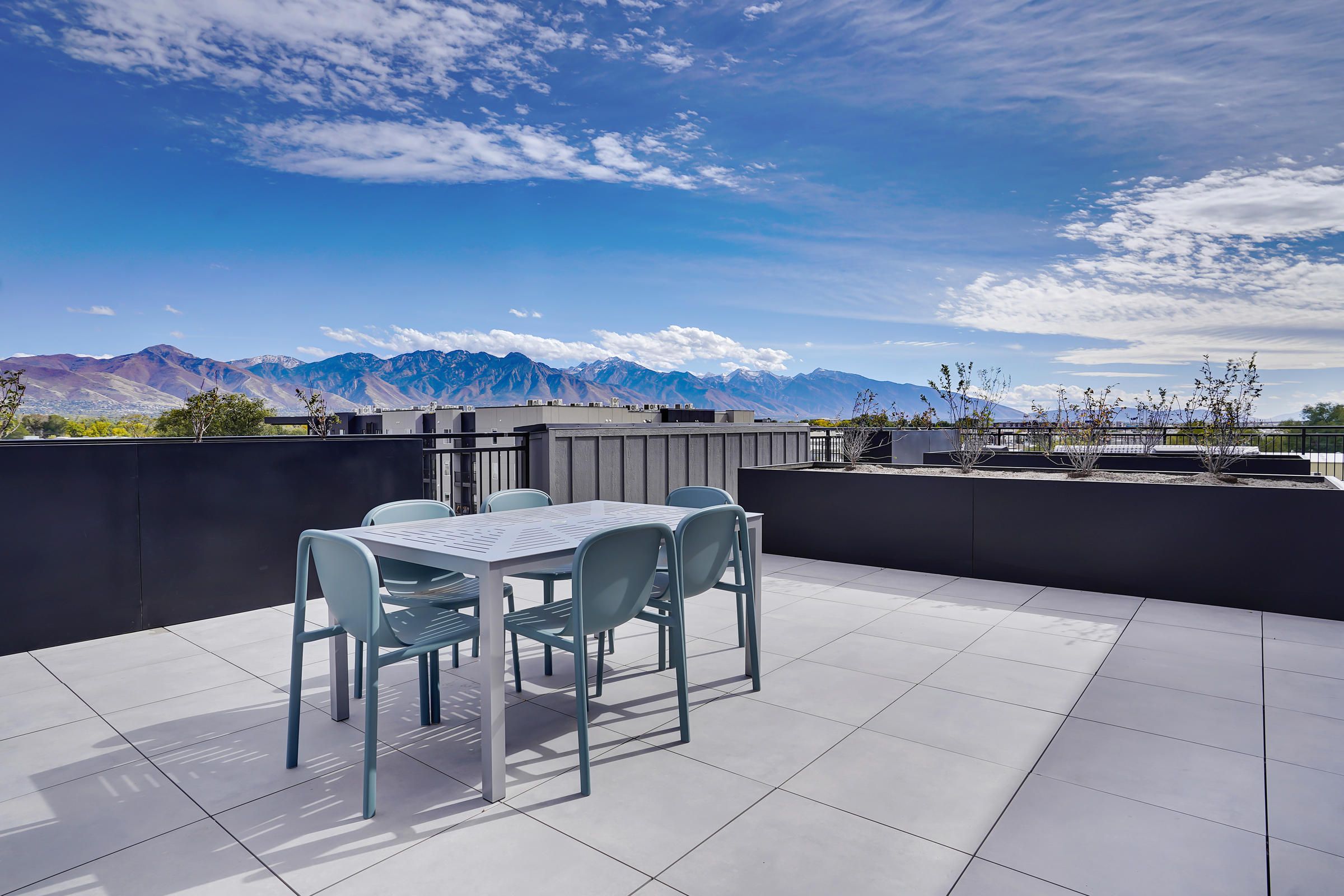Modern rooftop patio with a table and chairs, overlooking mountains under a blue sky with scattered clouds.