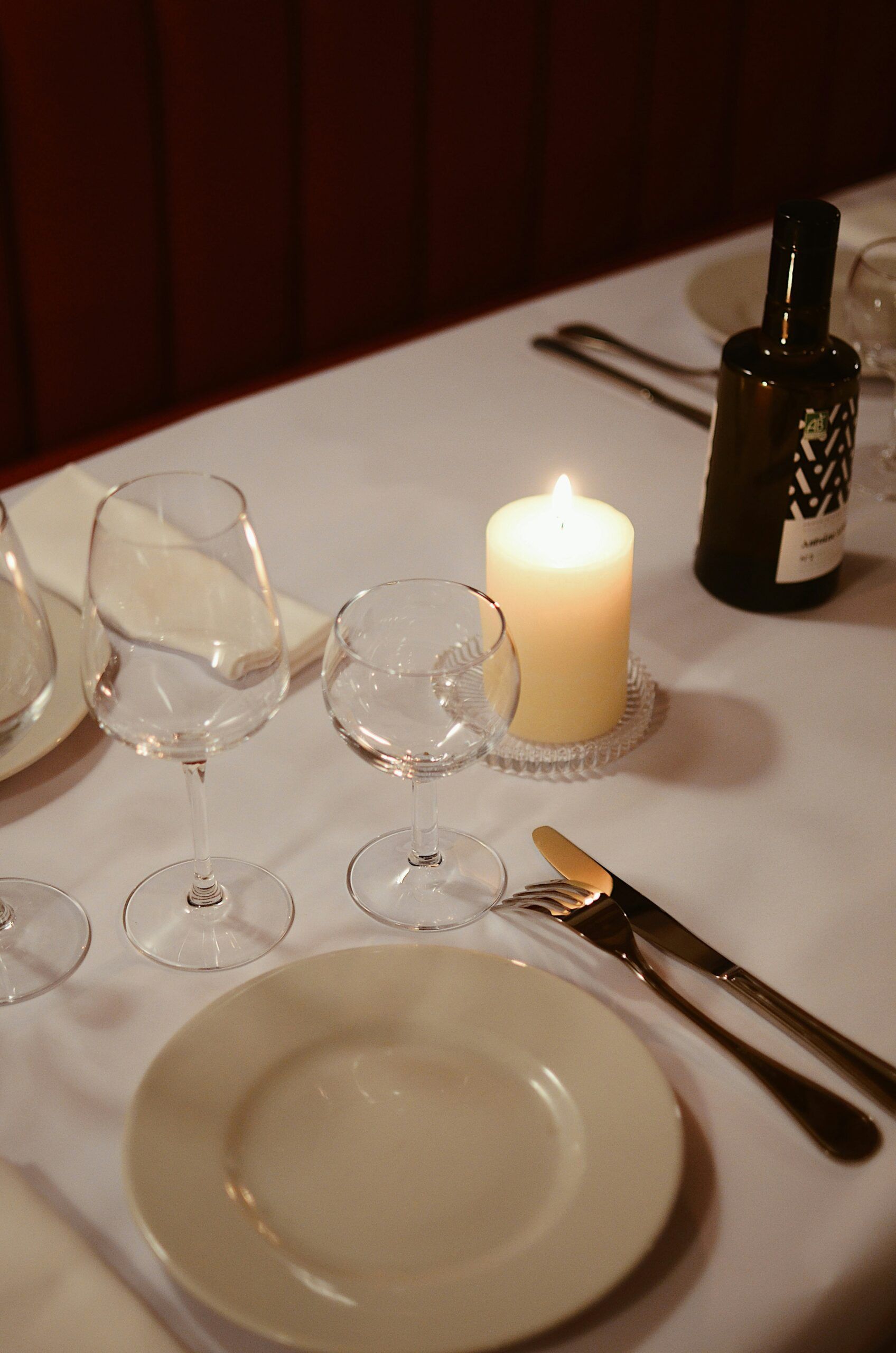 Elegant table setting with wine glasses, plates, cutlery, a lit candle, and a bottle on a white tablecloth.