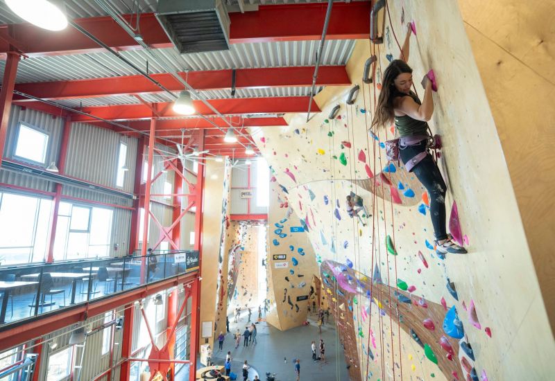 Woman climbing a tall indoor rock wall at a busy climbing gym with people below.