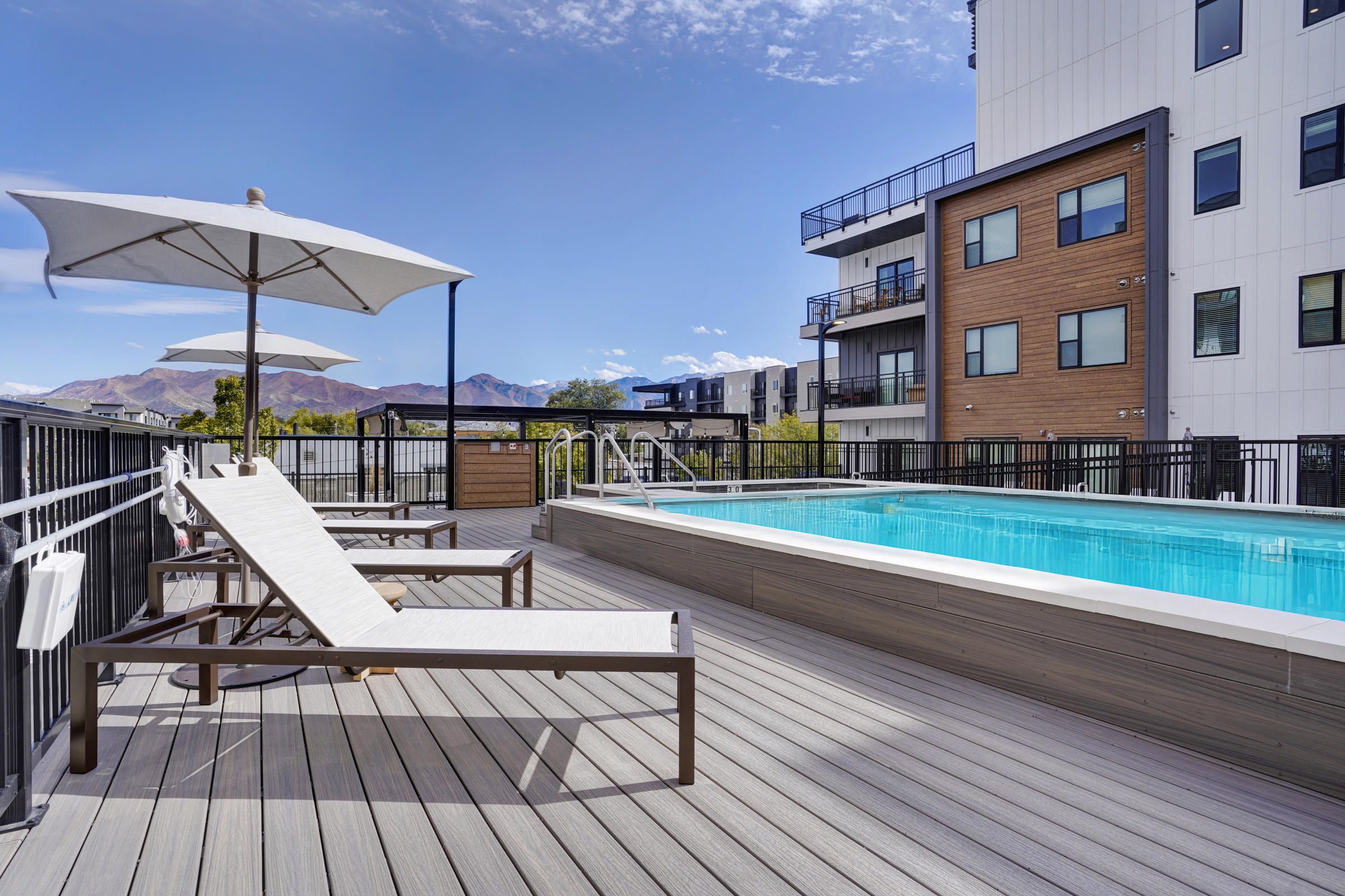 Modern apartment pool area with lounge chairs, umbrellas, and mountain views under a blue sky.