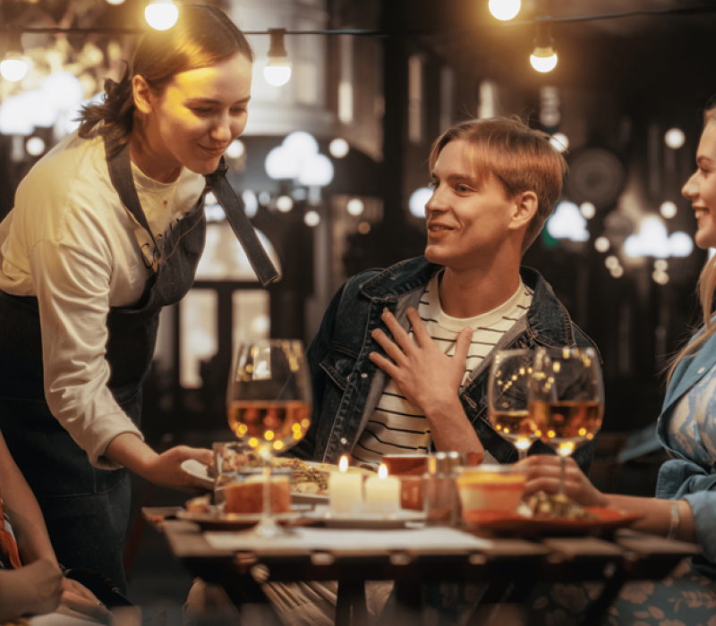 A waitress serves food to three smiling people at an outdoor restaurant table with wine and candles.