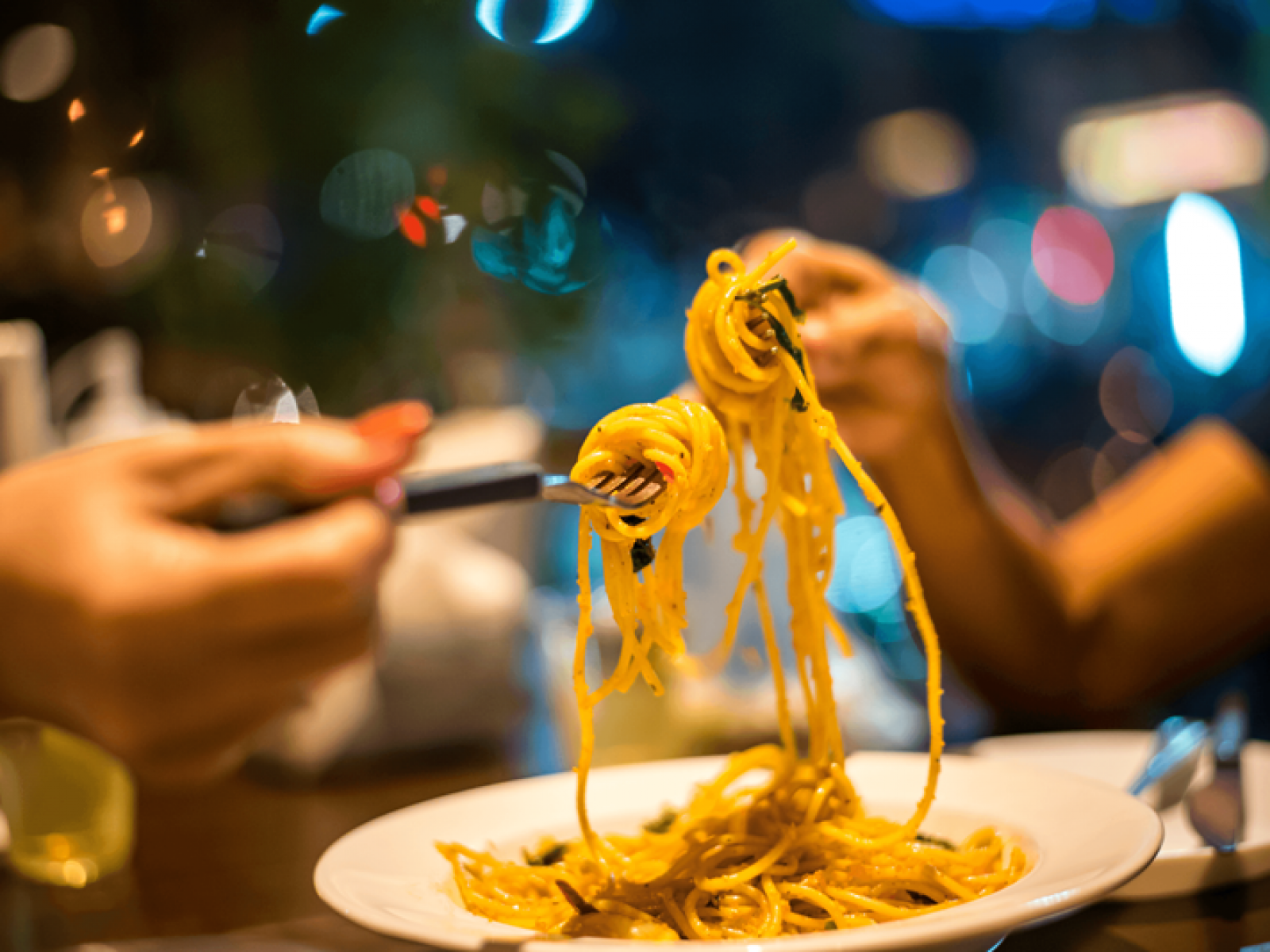 Two people twirl spaghetti with forks over a white plate in a warmly lit, blurred restaurant setting.