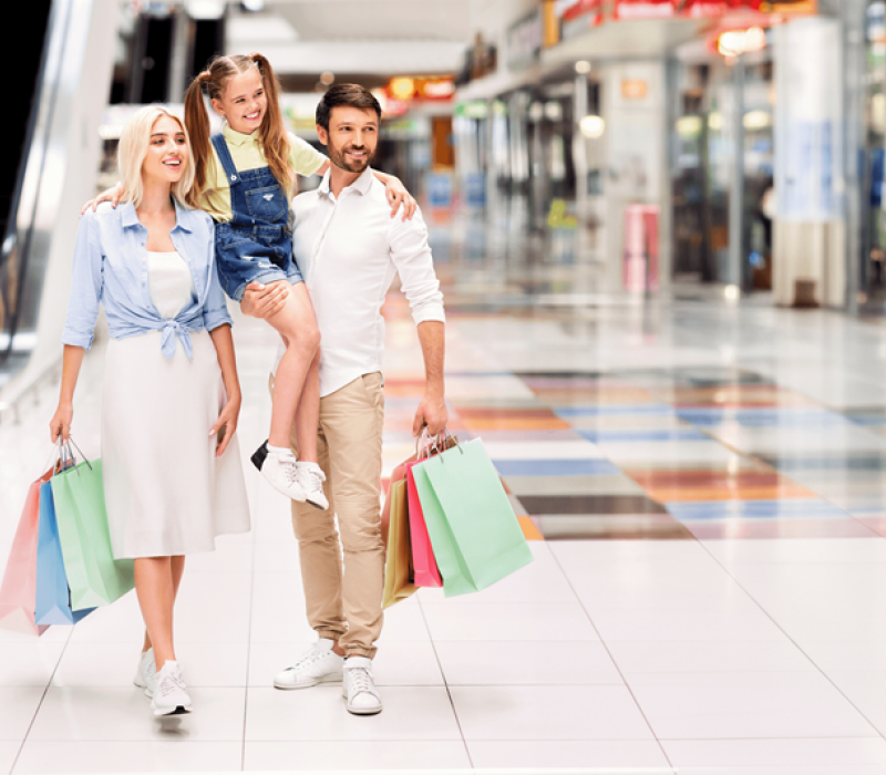 Family with shopping bags walks in a brightly lit mall; child rides on dad's back, all smiling.