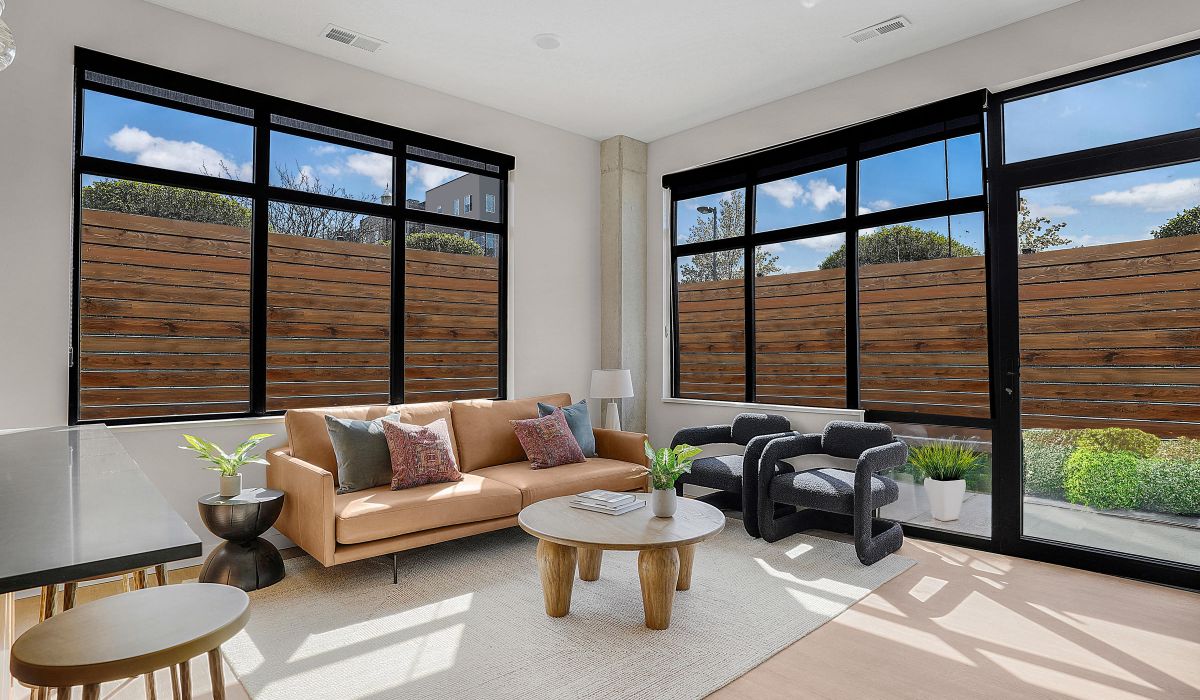 Modern living room with large windows, tan sofa, two black chairs, and a wooden coffee table.