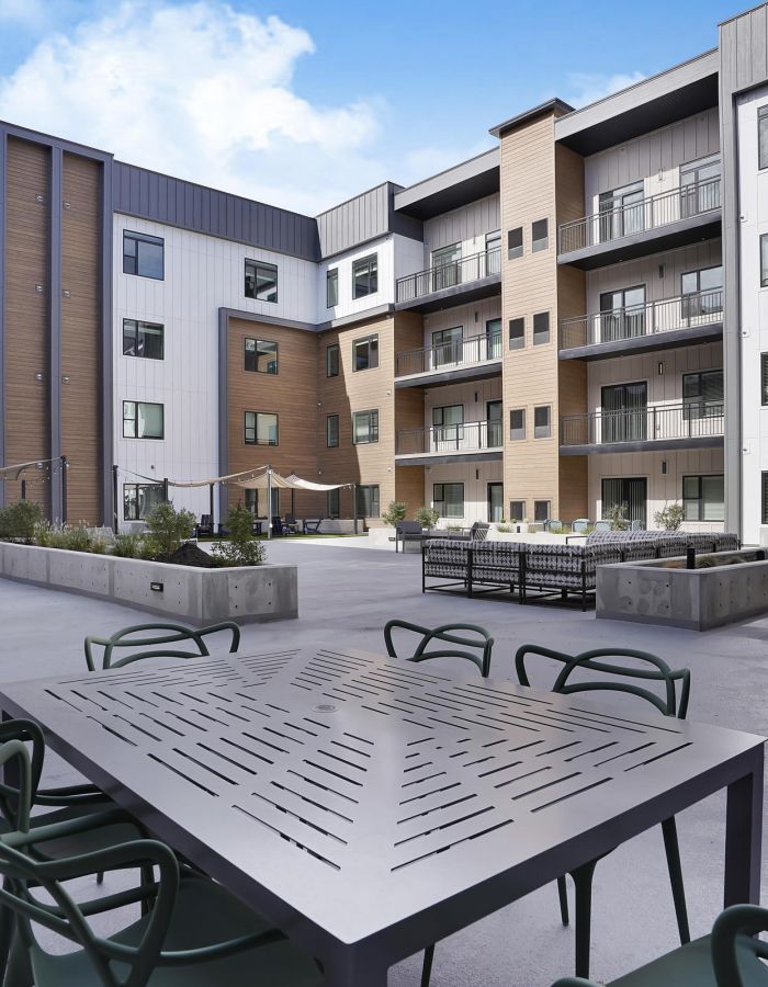 Modern apartment courtyard with outdoor seating, tables, planters, and multi-story buildings in the background.