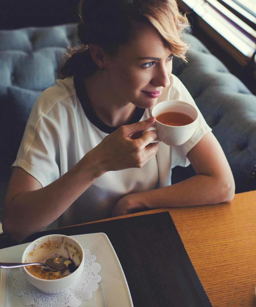 Woman sitting at a table, drinking tea, with a bowl of cereal and a teapot in front of her.