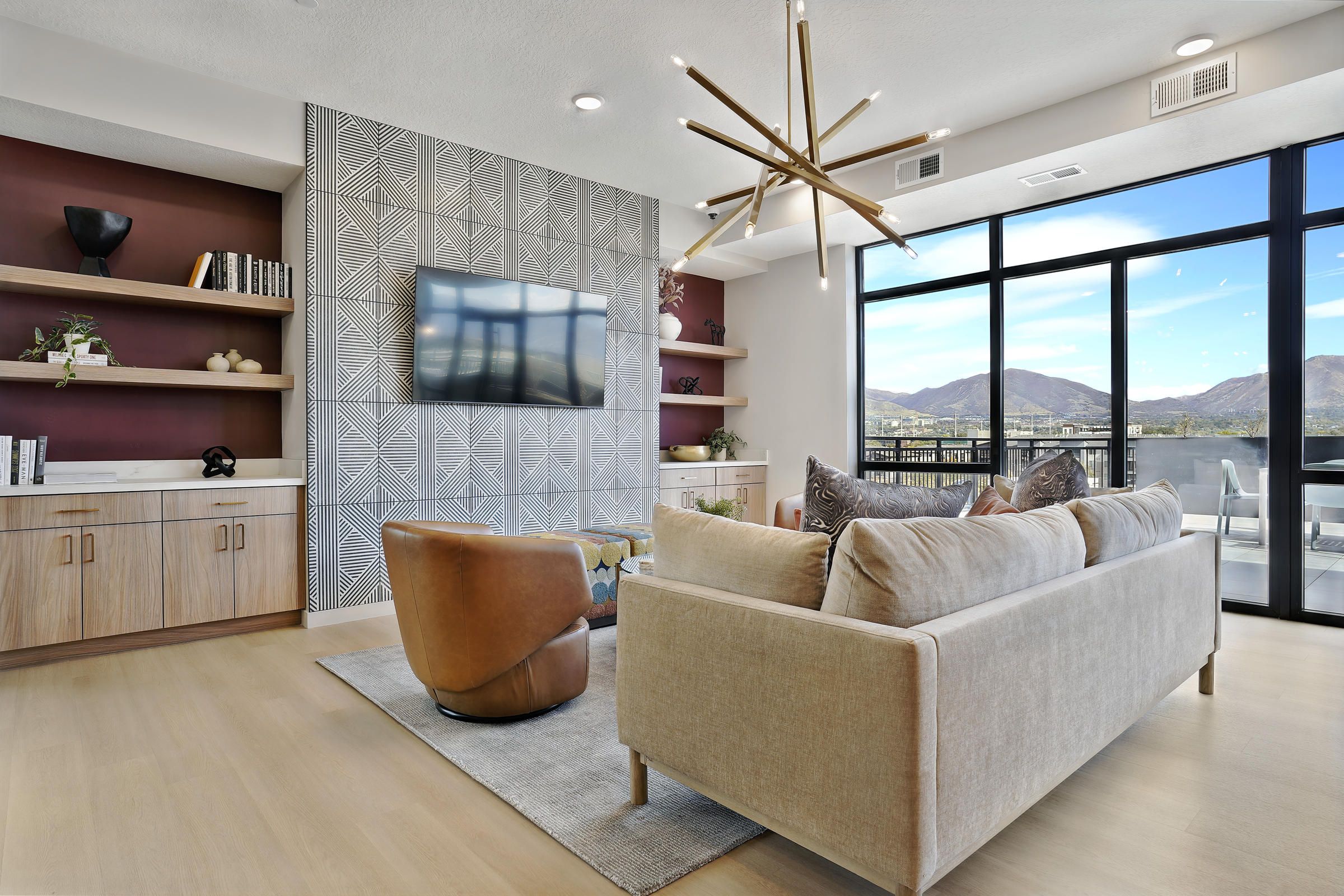 Modern living room with beige sofa, leather chair, TV, geometric accent wall, and large windows with mountain views.